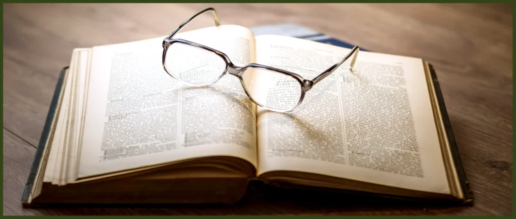 A wooden table with an open book laying on it with reading glasses resting on the pages of the book.