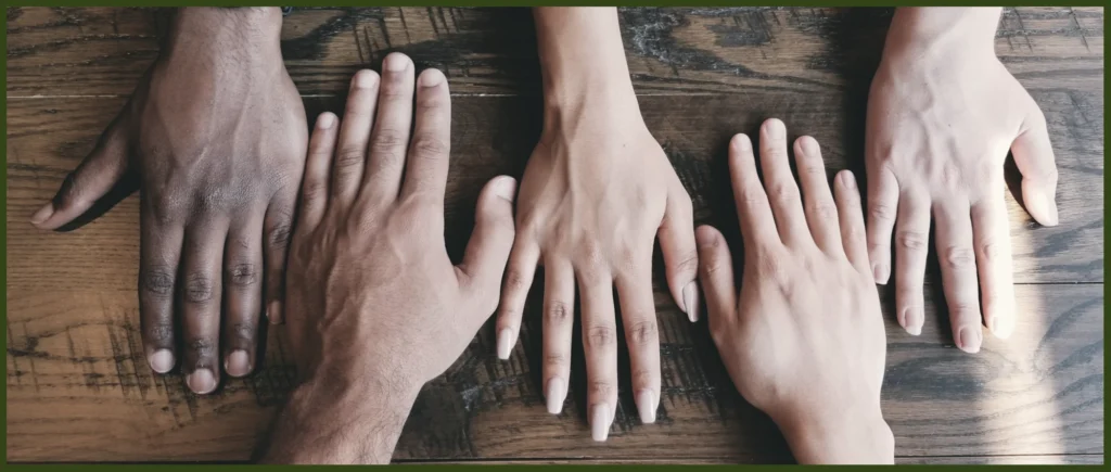 Five hands, with palms down, on a wooden table, representing cultural diversity and working together.